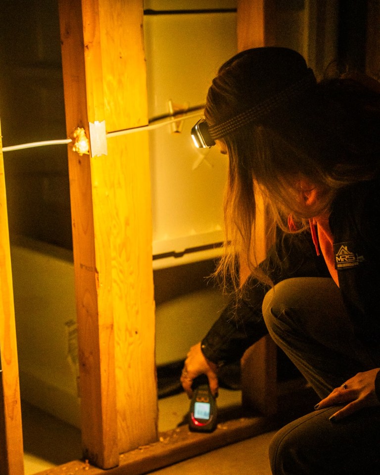 A technician in a headlamp crouches beside an open wall frame and takes a reading with a handheld meter.