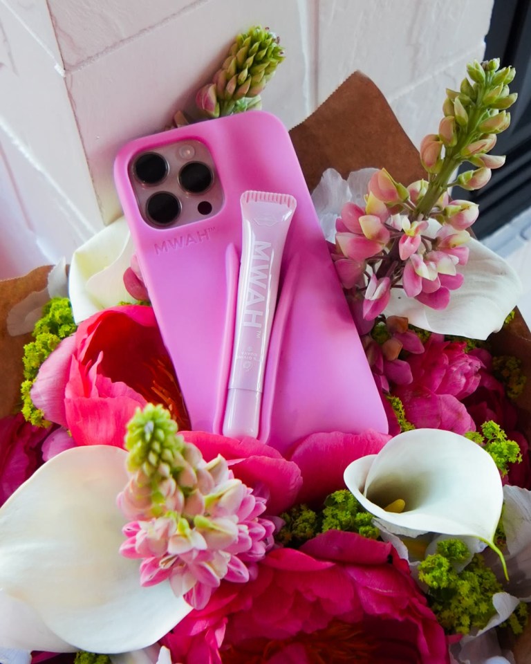 A pink MWAH phone case holding a matching MWAH tube, set in a bouquet of peonies, calla lilies and lupines.