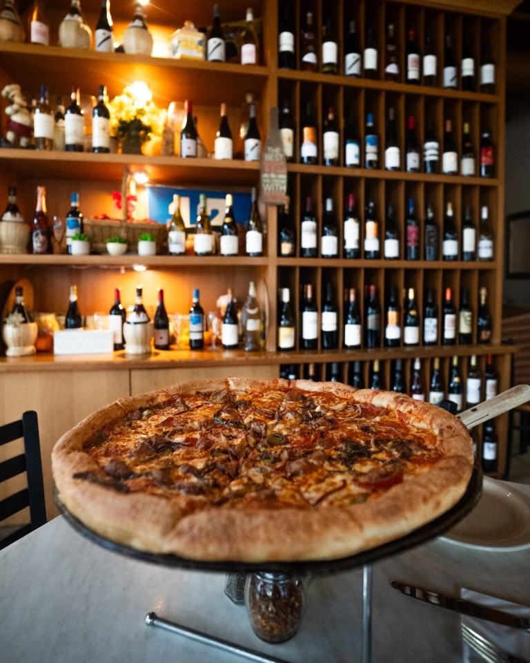 A loaded pizza on a raised serving stand in front of a warmly lit wall of wine bottles at Palermo's.