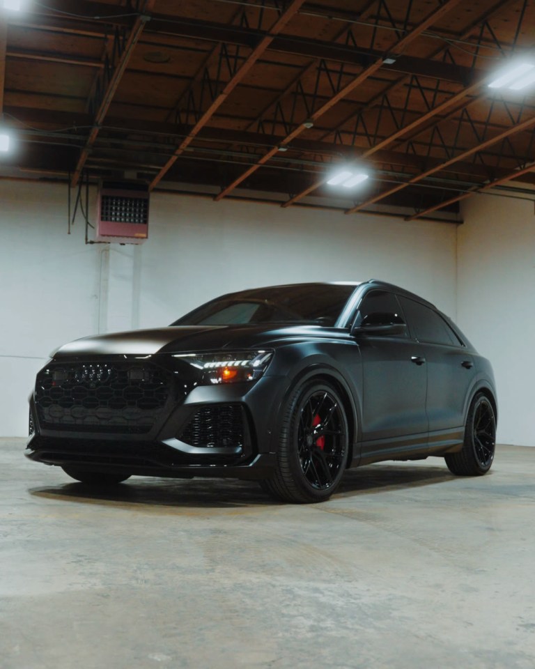 A satin black Audi SUV with red brake calipers parked in a warehouse under overhead lights.